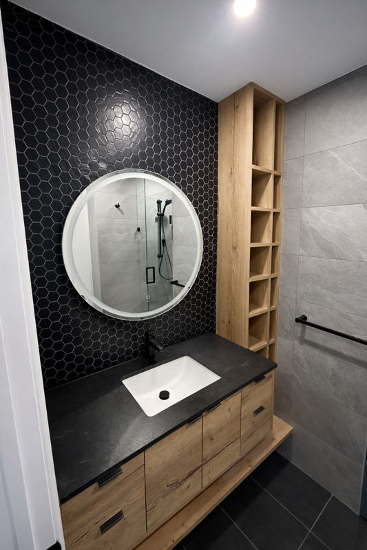 Bathroom vanity with a wood cabinet, black countertop, round mirror, and black hex tile accent wall.